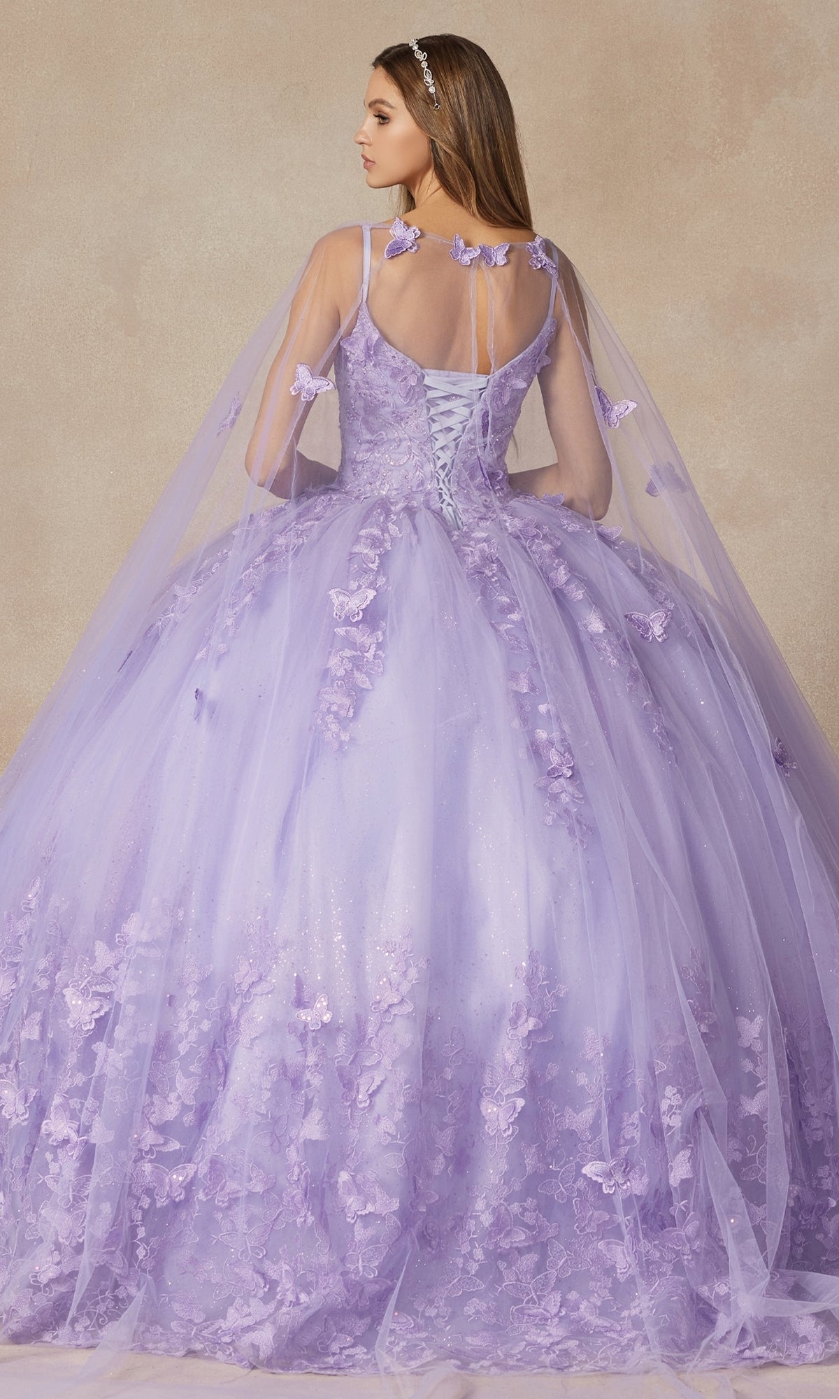 A woman in the Juliet 1442 Quinceañera Dress with Sheer Cape stands facing away, highlighting its elegant lace-up back, floral appliqués, and long, full tulle ball gown skirt.